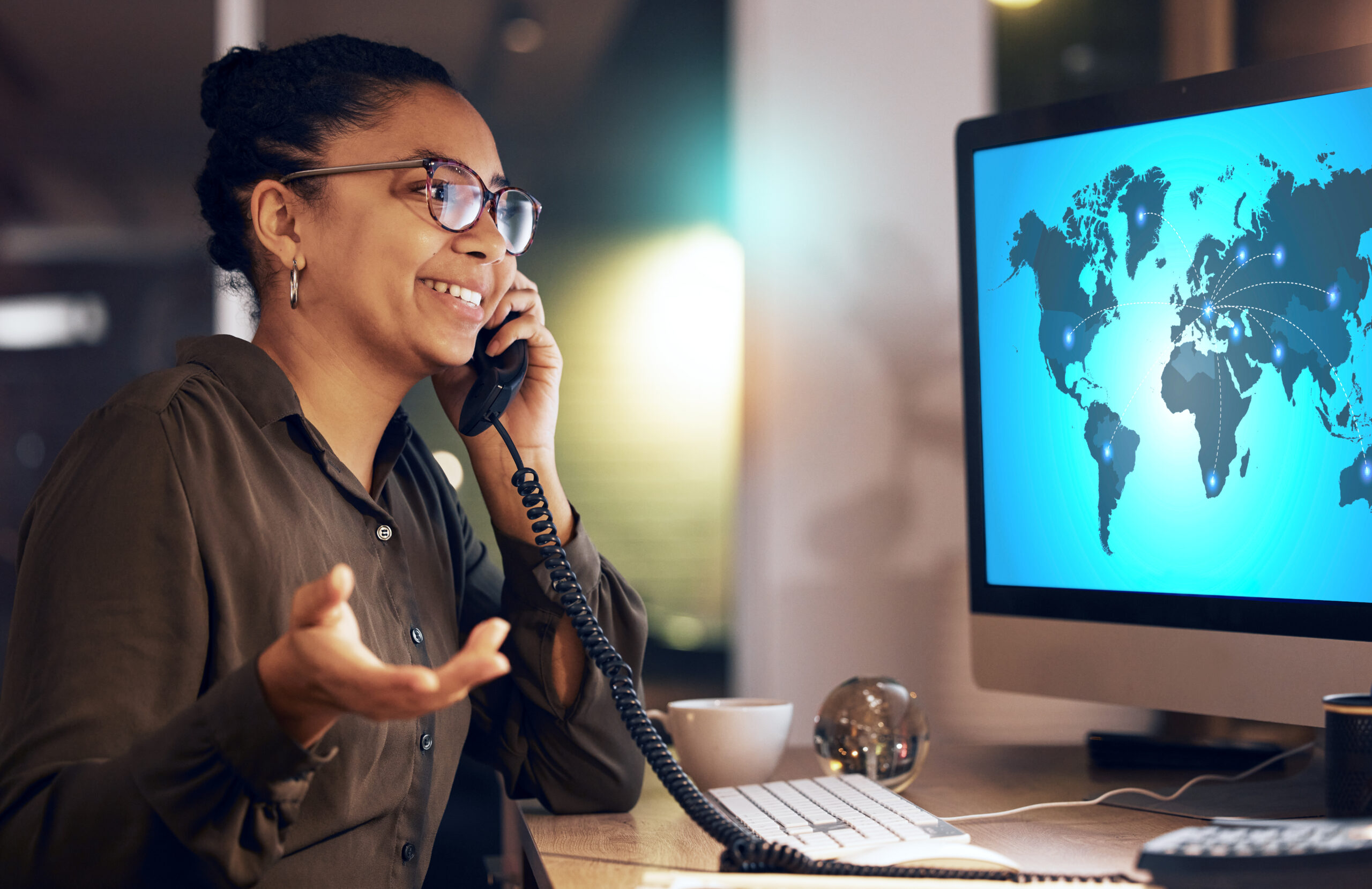 Businesswoman using an international VoIP service at her desk, speaking on a landline phone while viewing a digital world map on her computer screen.